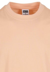 T-shirt à col rond de couleur pêche en tissu de coton doux, avec une étiquette affichant "Urban Classics" et la taille "M". Texture lisse, design minimal.