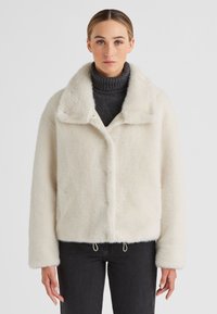 Weißes, flauschiges, kurz geschnittenes Jackett mit weitem Kragen, Druckknopfverschluss und seitlichen Kordelzügen, kombiniert mit einem dunklen Rollkragenpullover und schwarzen Jeans.