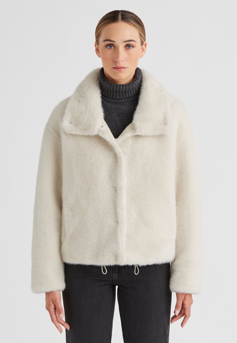 Weißes, flauschiges, kurz geschnittenes Jackett mit weitem Kragen, Druckknopfverschluss und seitlichen Kordelzügen, kombiniert mit einem dunklen Rollkragenpullover und schwarzen Jeans.