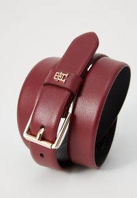 Ceinture en cuir rouge avec une boucle en ton or et un logo en relief. Elle se distingue par sa texture lisse et son design élégant, mettant en avant sa longueur ajustable.
