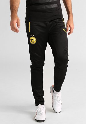 Mann trägt schwarze Puma-Trainingshose mit gelbem BVB 09-Logo und weißen Sportschuhen, läuft auf schlichtem hellem Hintergrund.