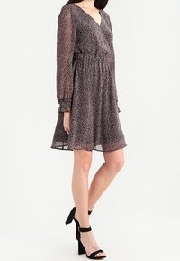 Vestido de manga larga, con un estampado oscuro, escote en V, cintura ajustada y falda fluida. Se complementa con sandalias negras de tacón alto.