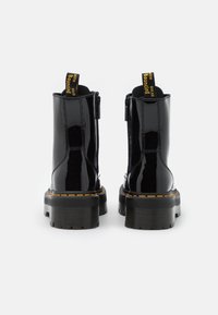 Botas de tobillo de charol negro con suela gruesa, costuras amarillas y lengüetas en la parte trasera. Cierre de cremallera en el lateral.