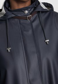 Navy wodoodporna kurtka z regulowanym kapturem, srebrnymi elementami i kontrastującymi szarymi akcentami. Posiada regulowane sznurki oraz zatrzaski.