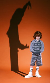 Felpa grigia con motivi audaci, pantaloni corti abbinati, scarpe alte bianche e calze con lettering, su uno sfondo arancione.