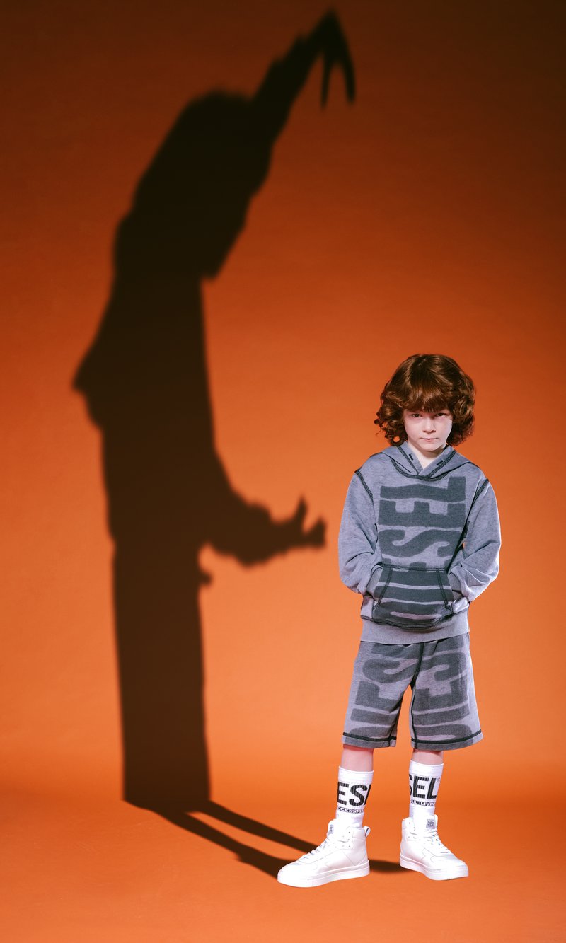Felpa grigia con motivi audaci, pantaloni corti abbinati, scarpe alte bianche e calze con lettering, su uno sfondo arancione.
