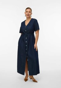 Mujer con un vestido largo azul oscuro abotonado, de mangas cortas y cinturón anudado en la cintura, combinado con tacones slingback marrones de punta puntiaguda.