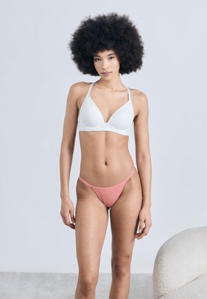 Femme aux cheveux afro portant un soutien-gorge blanc et un slip bikini rose, debout devant un fond clair uni près d'une chaise texturée.