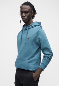 Kapuce ar kapuci izgatavots no tirkīza kokvilnas, ar priekšējo kabatu un ribveida aprocēm. Mazs dzeltens logotips izšūts uz krūtīm.