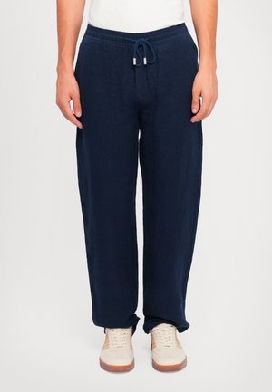 Pantalons en lin bleu marine avec une taille élastique et un cordon de serrage, présentant une coupe ample et un devant plat, associés à des baskets beiges.