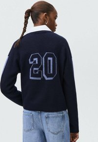 Mornarska pletena puloverja z modro številko "20" na hrbtu, dolgimi rokavi in sproščenim krojem. Nosi se z svetlim prati kavbojkami.