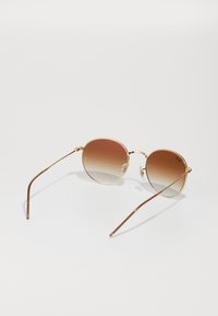 Ochelari de soare din metal aurit cu lentile rotunde, având un nuanțat de căldură maronie, brațe subțiri și tampoane pentru nas transparente. Design minimalist și ușori.