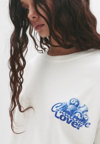 Bílá bavlněná košile s dlouhým rukávem, na které je modrý grafický potisk s válečky a textem „Challenge Love.“ Hladká textura, kulatý výstřih.