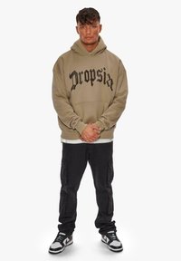 Übergroßer beiger Hoodie mit schwarzem Text, Kängurutasche; kombiniert mit dunklen Cargo-Hosen und schwarz-weißen Sneakern. Einfarbiger Hintergrund.
