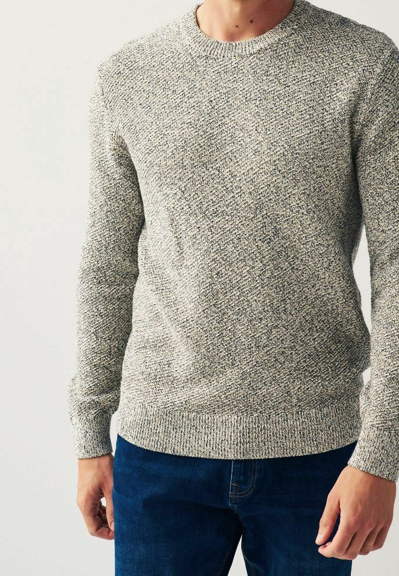 Homme portant un pull en maille texturée crème et gris avec poignets et ourlet côtelés, associé à un jean en denim bleu foncé.