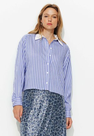 Chemise à rayures bleu clair avec manches longues et col blanc, portée par-dessus une jupe bleu pailletée. Coupe courte.