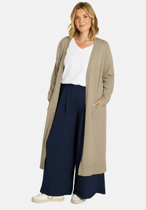 Femme portant un long cardigan beige, un top blanc en V, un pantalon large bleu marine et des baskets blanches, debout avec une main dans la poche.