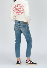 Biała kurtka bomber z czerwoną logo, niebieskie wąskie dżinsy z haftowanymi tylnymi kieszeniami oraz czarno-czerwone sneakersy z kremowymi podeszwami.