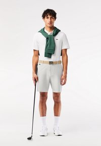 Homme en polo blanc et short avec un pull vert sur les épaules tenant un club de golf, debout sur un fond blanc uni.