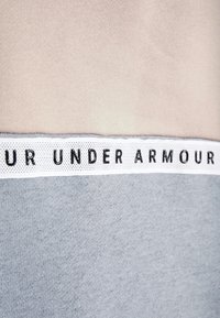 Crewneck-trui met beige, lichtgrijze en donkergrijze panelen. Een witte band met perforaties toont de tekst "UNDER ARMOUR" in zwarte letters.