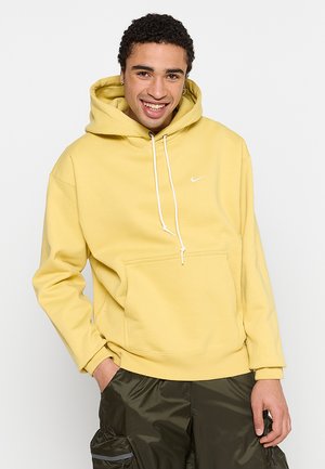 Gul bomulds hoodie med en frontlomme, hvide snørebånd og et lille hvidt logo på brystet. Stoffet har en glat tekstur.