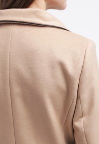 Vrouw in een beige wollen jas met zwarte bies langs de kraag, van schouder tot nek afgebeeld tegen een effen achtergrond.