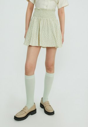 Falda verde claro con cintura fruncida, que presenta un pequeño patrón floral blanco. Combinada con calcetas verde menta hasta la rodilla y mocasines beige.