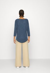Blau geflecktes Strickoberteil mit Dreiviertelärmeln, abgerundetem Saum, kombiniert mit hoch taillierten beigen weiten Hosen und weißen Sneakers.