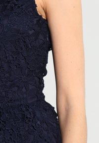 Abito senza maniche blu navy con dettagli in pizzo floreale intricati, superficie testurizzata e tessuto liscio attorno al collo e alle aperture per le braccia.