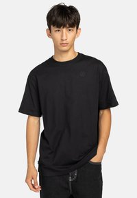 Zwarte katoenen t-shirt met ronde hals, korte mouwen en een subtiele logopatch op de borst. Heeft een losse pasvorm en een natuurlijke drapering.