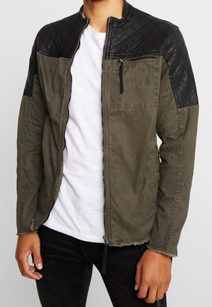 Homme portant une veste verte dézippée avec des épaules matelassées noires par-dessus un t-shirt blanc et un pantalon noir.