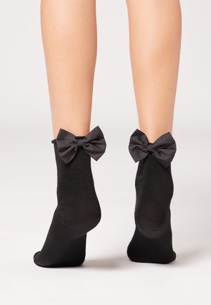 Schwarze Knöchelsocken aus weichem Stoff, mit großen schwarzen Satinschleifen an den Bündchen, die ein einzigartiges und strukturiertes Detail bieten.