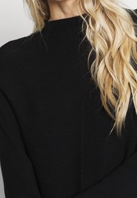 Femme blonde aux cheveux ondulés portant un pull à col roulé noir texturé, visible des épaules au cou.