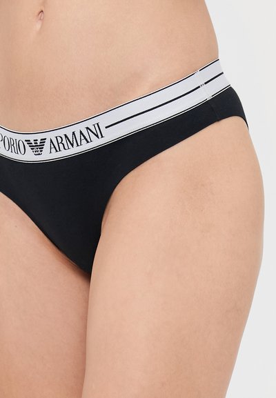 Black thong with a soft, stretchy fabric; features a white elastic waistband with black "Emporio Armani" branding and two thin stripes.