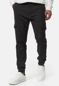 Pantalones cargo negros fabricados con un material suave, con una cinturilla elástica, puños acanalados y bolsillos laterales. Llevados con zapatillas blancas.