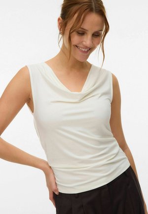 Kvinde med rødt hår iført en ærmeløs hvid top med draperet hals og sorte bukser, smilende og poserende med den ene hånd på hoften.