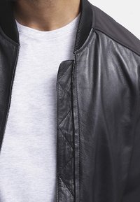 Černá kožená bomber bunda s žebrovaným límcem a prošívanými detaily podél zipu, přeložená přes světle šedé tričko.