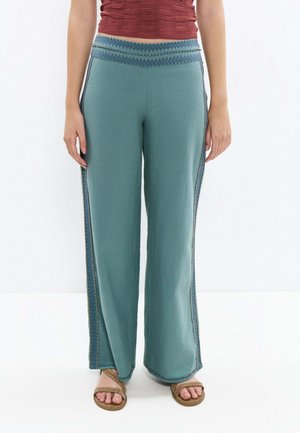 Personne portant un pantalon large bleu sarcelle avec une taille à motif et des bandes latérales, associé à des sandales marron cloutées et un top court rouge.