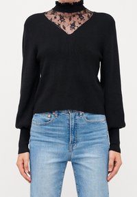 Maglione nero lavorato a maglia con scollo a V e sovrapposizione di pizzo nero trasparente sul collo. Maniche lunghe a sbuffo; indossato con jeans in denim blu chiaro.