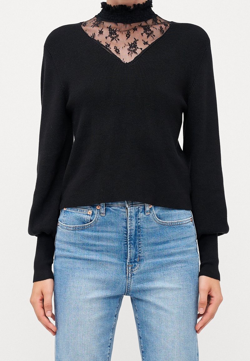 Maglione nero lavorato a maglia con scollo a V e sovrapposizione di pizzo nero trasparente sul collo. Maniche lunghe a sbuffo; indossato con jeans in denim blu chiaro.