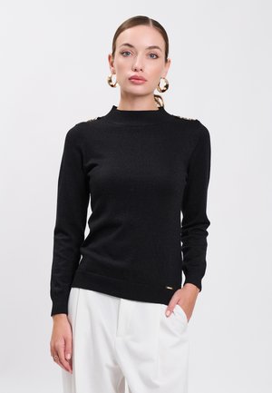 Femme portant un pull en maille noir à manches longues avec des boutons dorés sur les épaules, de grandes boucles d’oreilles dorées en forme d’anneau et un pantalon blanc, debout devant un fond blanc.