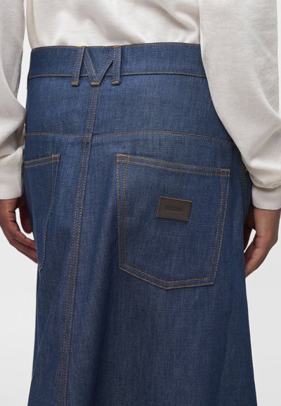 Gros plan d'une personne portant une jupe en denim bleue avec des poches arrière et un haut blanc à manches longues, montrant des détails de couture jaunes visibles.