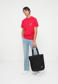 Mladý muž v červenom tričku, svetlo modrých džínsach a bielych teniskách, ktorý drží čiernu tašku značky Tommy Jeans pred bielym pozadím.