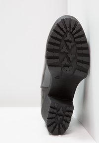 Černá podrážka boty s texturovaným gumovým designem a hlubokým běhounem pro lepší trakci, kulatým tvarem a hladkým povrchem na horní straně.