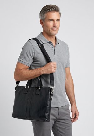 Man draagt een grijze polo en broek en houdt een zwarte leren schoudertas met verstelbare riem tegen een lichte achtergrond.