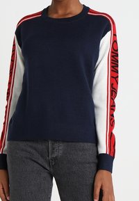 Pull en tricot bleu marine avec des manches rayées rouges et blanches, arborant le logo "TOMMY JEANS" en rouge. Col rond et coupe décontractée.