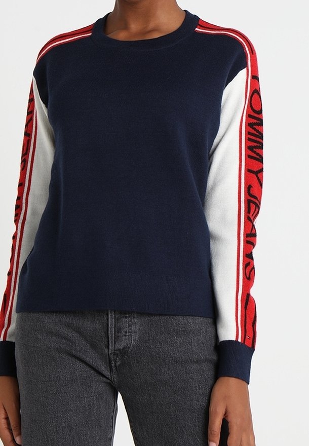 Pull en tricot bleu marine avec des manches rayées rouges et blanches, arborant le logo "TOMMY JEANS" en rouge. Col rond et coupe décontractée.