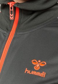 Svart idrottsjacka med hög krage, orange dragkedja och broderad "hummel"-logotyp i orange på vänster sida. Slätt tyg.