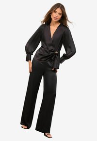 Czarny satynowy top zapinany na zakładkę z głębokim dekoltem w kształcie litery V i długimi balonowymi rękawami, stylizowany z węzłowym detalem oraz szerokimi spodniami z tej samej tkaniny.