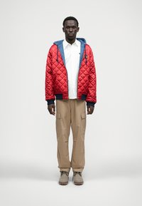 Κόκκινο καπιτονέ μπουφάν με μπλε επένδυση, λευκό πουκάμισο από μέσα, μπεζ παντελόνι cargo και γκρι μπότες. Το μπουφάν διαθέτει κλείσιμο με φερμουάρ και κορδόνια προσαρμογής.